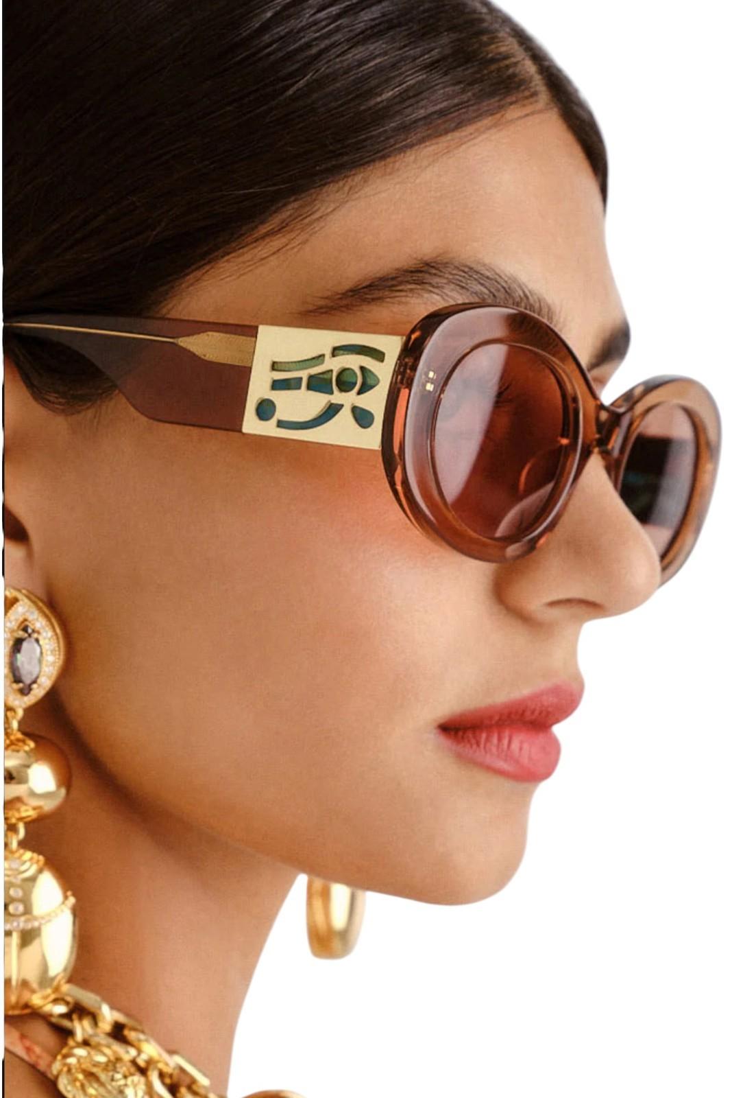 Close-up of a woman wearing sunglasses with a brand logo, gold earrings, and a gold necklace.