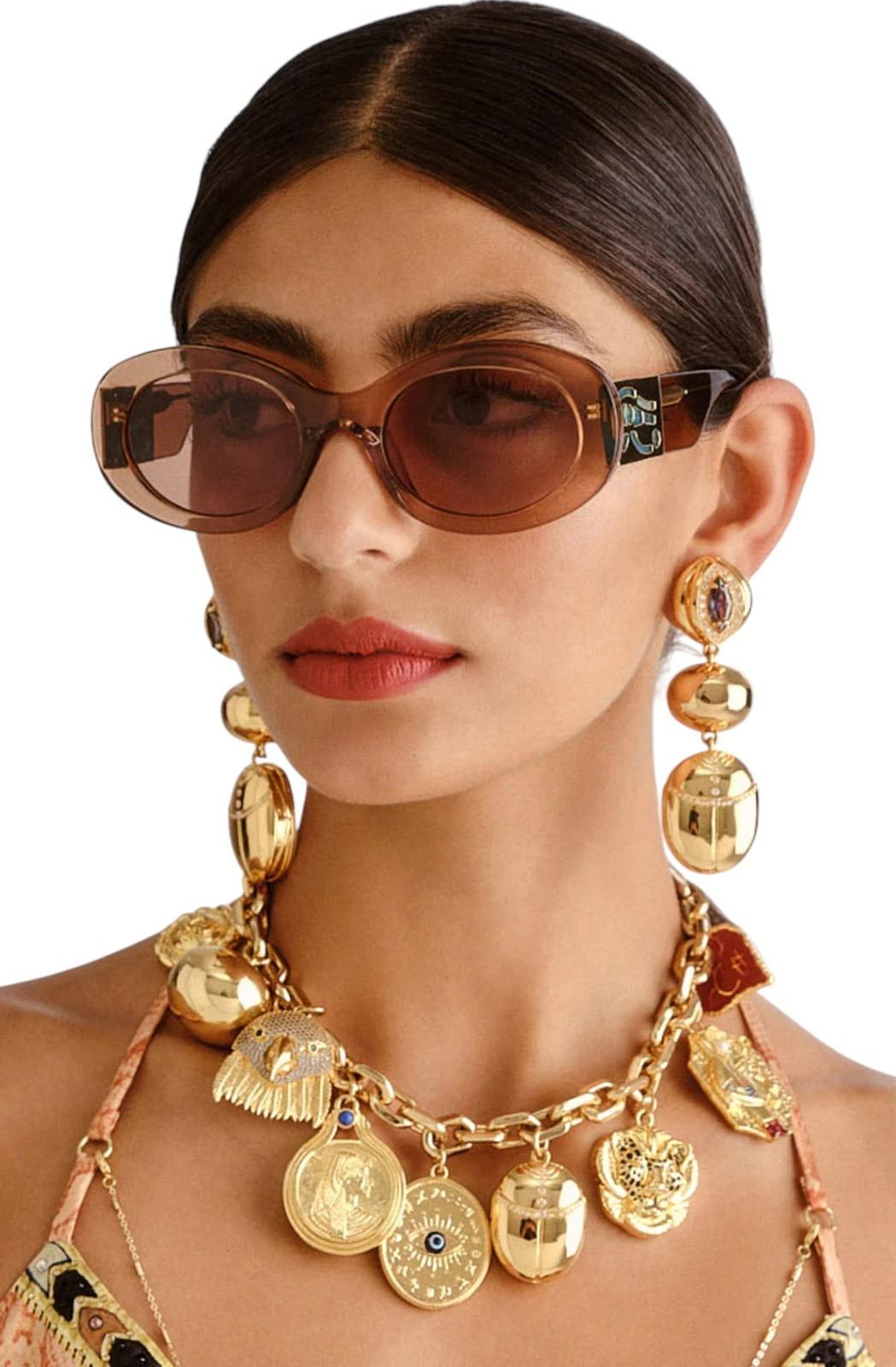 Woman wearing gold jewelry including earrings, necklace, and sunglasses on a white background