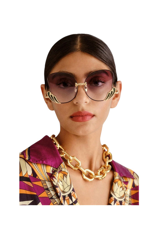 A model wearing oversized cat-eye sunglasses with gold metal frames and dark gradient lenses, paired with a chunky gold chain necklace and a patterned purple shirt, set against a plain white background.