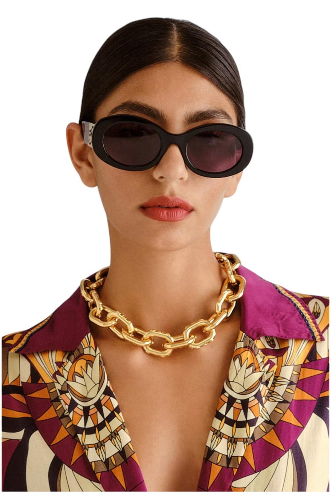 Woman wearing sunglasses and a patterned shirt with a gold chain necklace on a white background
