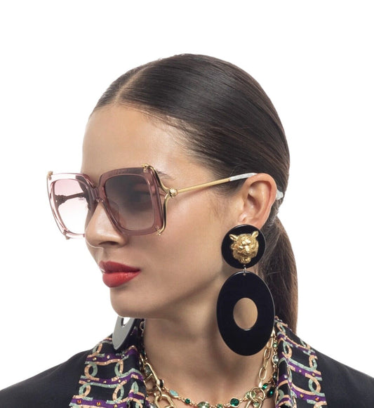 A woman wearing oversized square sunglasses with pink frames and gold metal details. The sunglasses have a vintage vibe and are designed by Camilla.