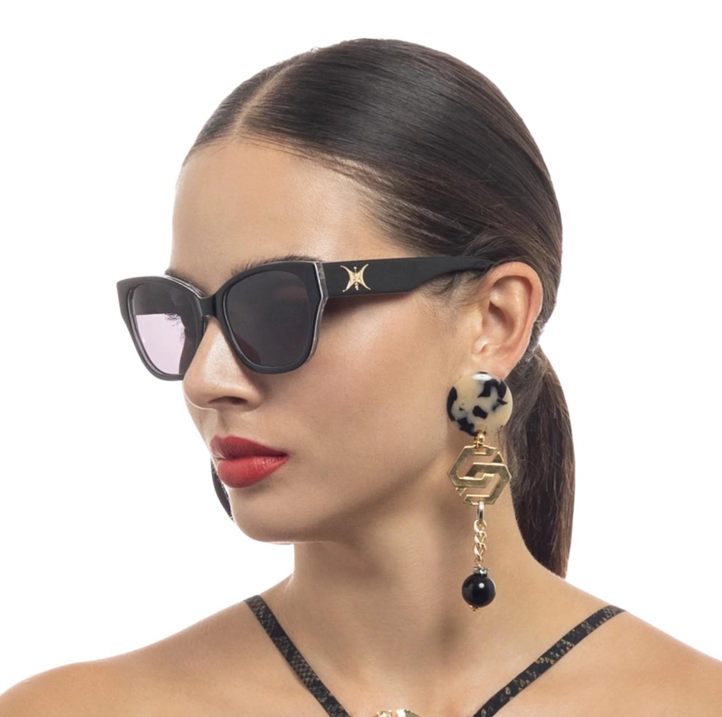 Woman wearing black sunglasses and a gold earring with a black bead on a white background
