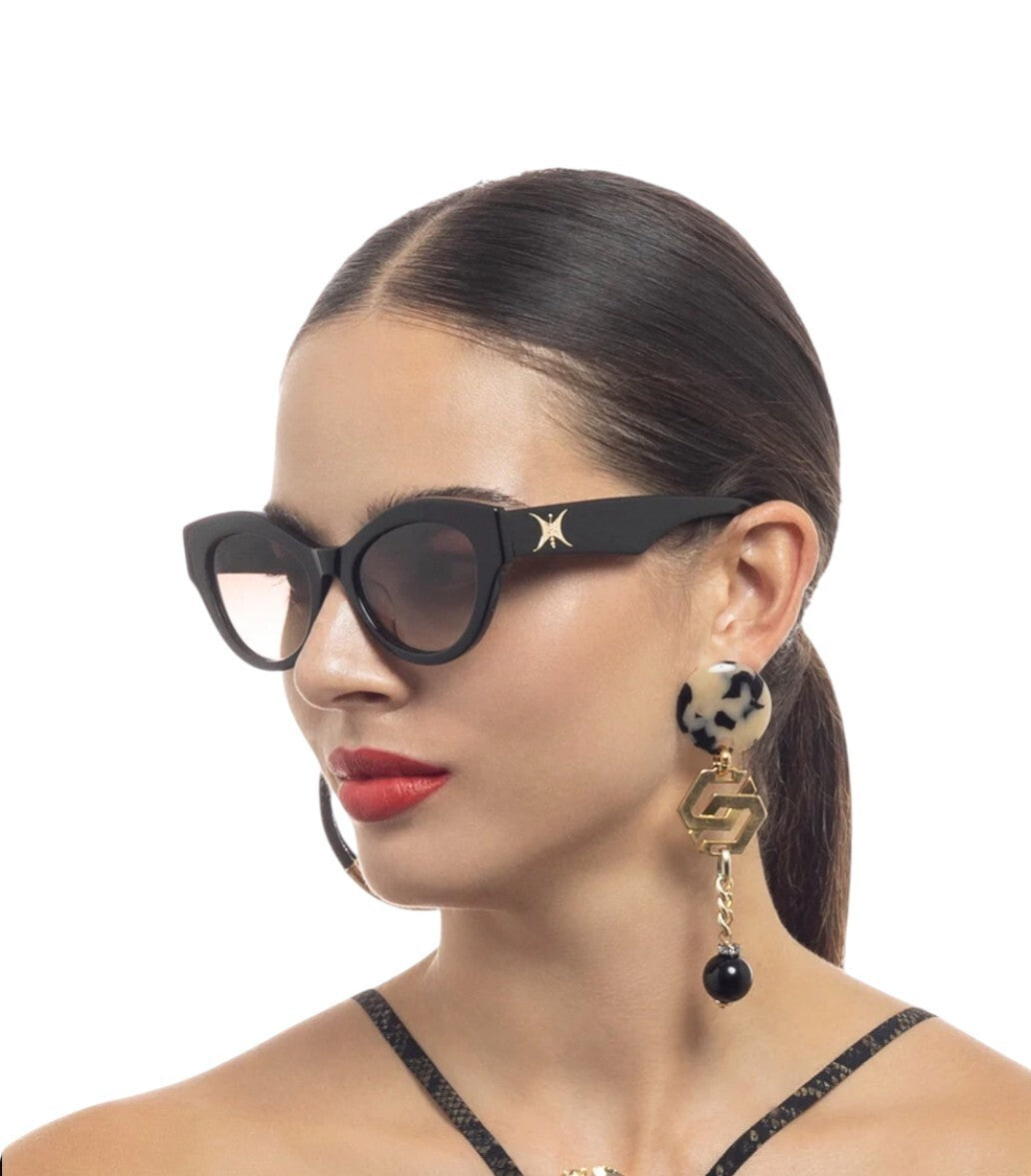 Woman wearing black cat-eye sunglasses and a dangling earring with a black bead on a white background
