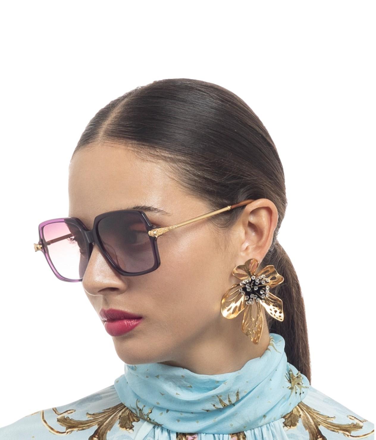 Woman wearing sunglasses and a light blue scarf with gold patterns on a white background