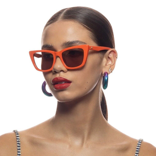 A woman wearing tangerine orange, cat eye shaped sunglasses with a medium size fit. She has red lipstick on and is accessorized with multiple earrings.