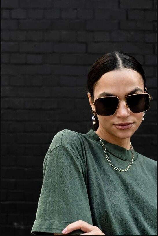 Naked Pear model wearing oversized gold metal Karen Walker Nada sunglasses with green mono lenses against a black wall