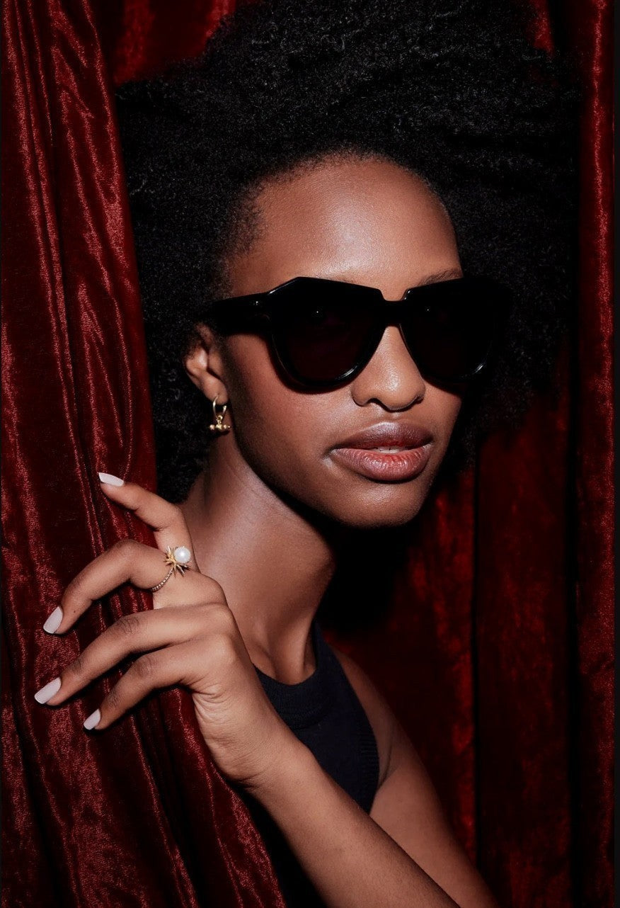 Model wearing black bio-acetate cat-eye Karen Walker Number One sunglasses with grey smoke lenses against red velvet