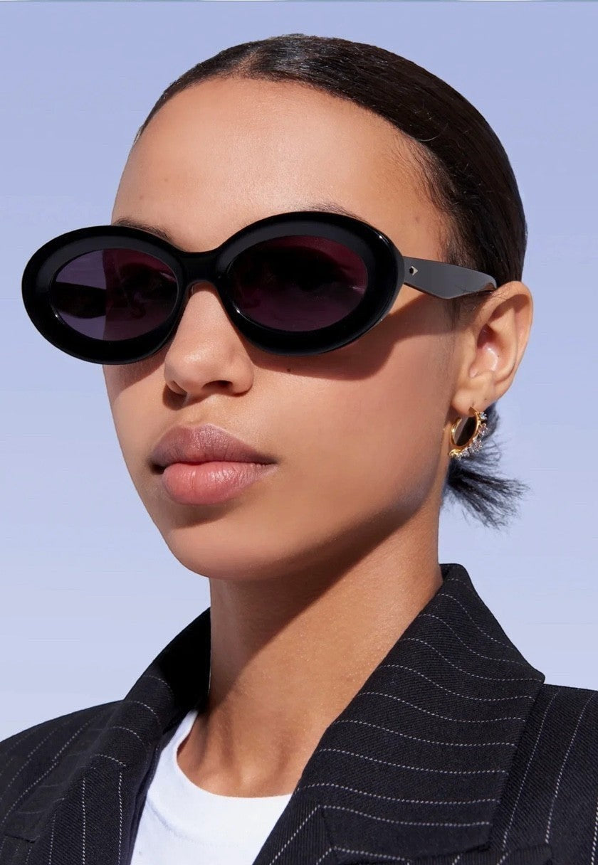 Model wearing Naked Pear Karen Walker Rapture B oval black bio-acetate sunglasses with grey lenses
