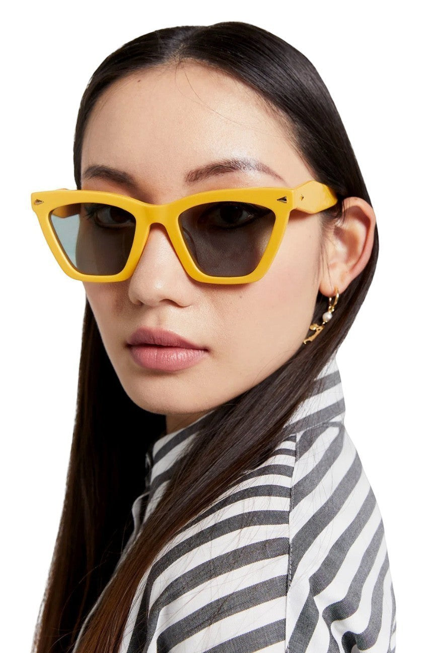 Model wearing Naked Pear Karen Walker Spellbound cat-eye sunglasses in marigold yellow acetate with emerald lenses.