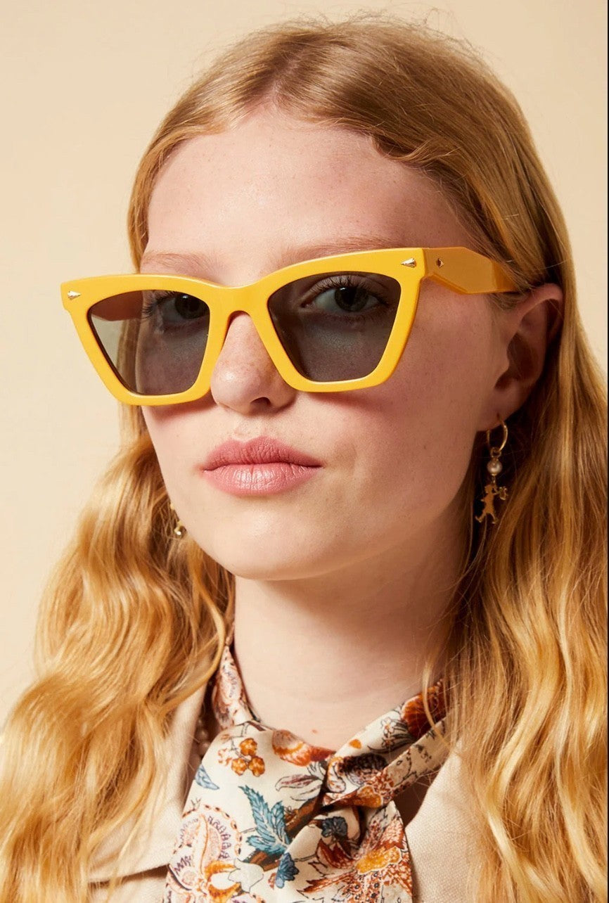 Model wearing Naked Pear Karen Walker Spellbound cat-eye sunglasses in marigold acetate with emerald-tinted lenses.