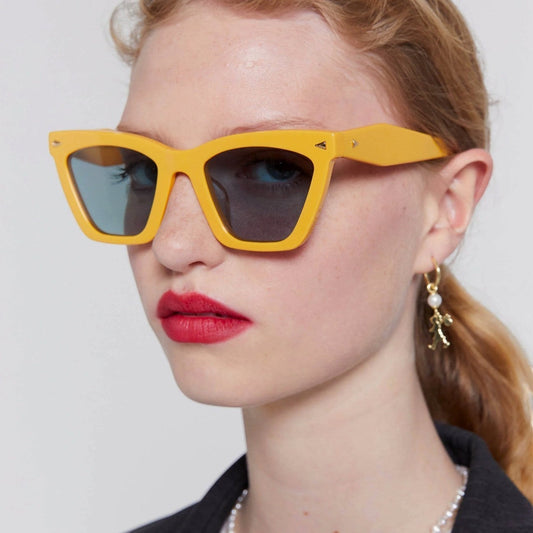 Model wearing Naked Pear Karen Walker Spellbound cat-eye sunglasses in glossy marigold acetate with emerald lenses.