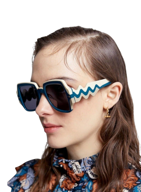A close-up of a woman wearing distinctive square sunglasses with dark lenses, cream and blue frames, and wavy blue detailing on the temple arm.