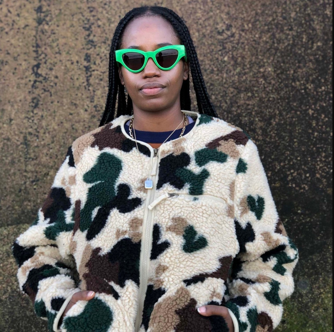 Person wearing a camouflage jacket and green sunglasses against a textured wall.
