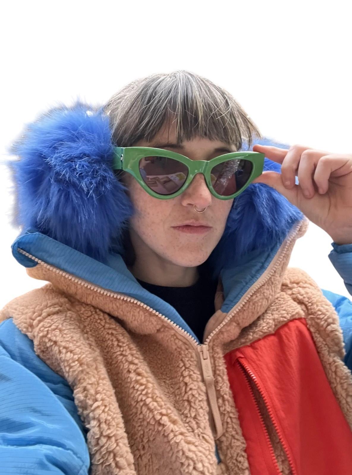 Person wearing a colorful jacket with a blue hood and red lining, holding green sunglasses.
