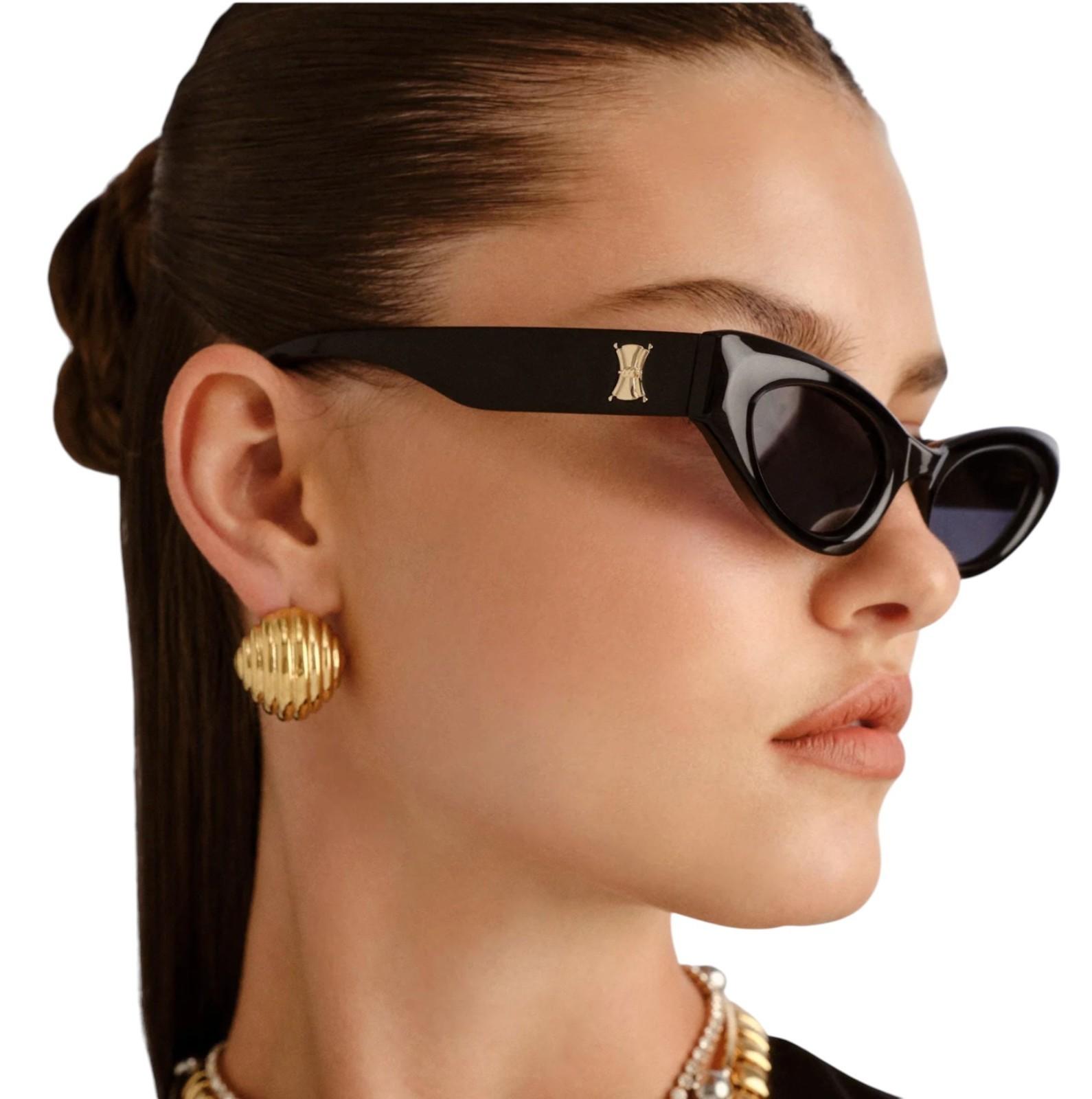 Woman wearing black sunglasses with a gold logo and gold earrings on a white background
