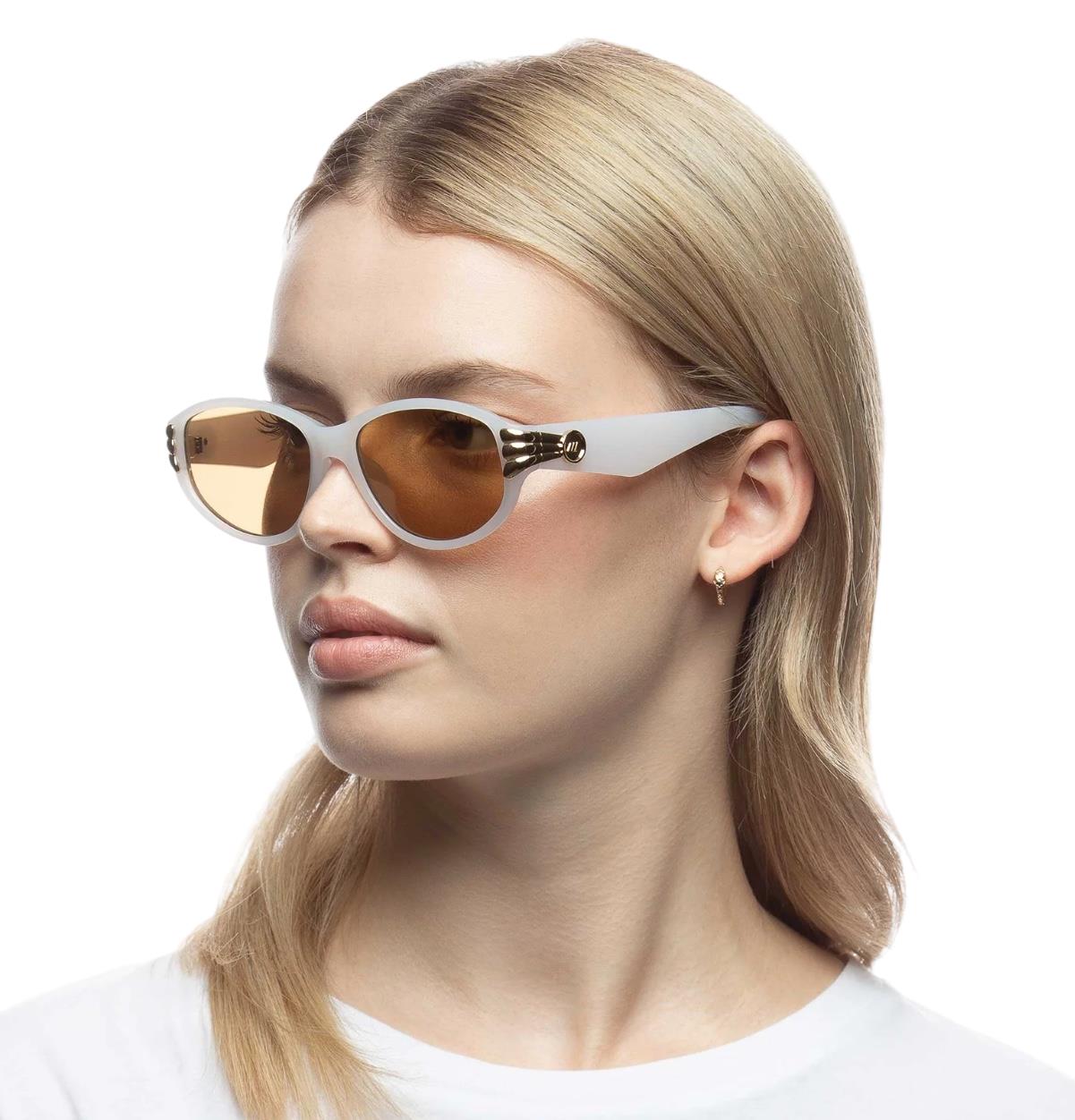 A model wearing white and gold Le Specs sunglasses with an oval shape and acetate material.