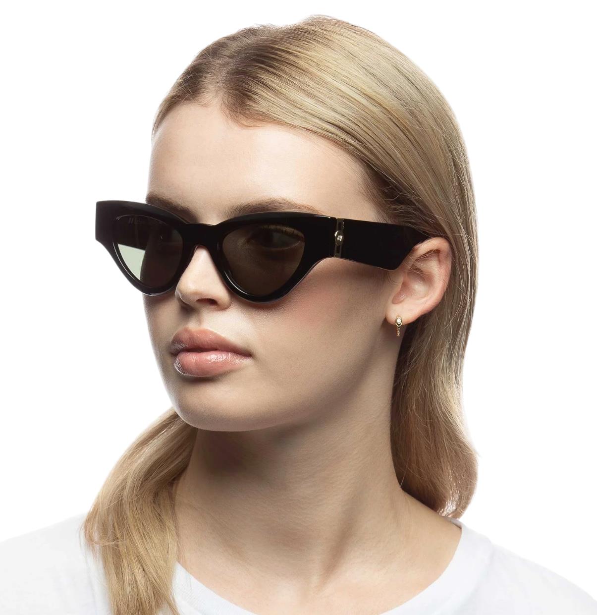 A woman wearing black Le Specs Fanplastico sunglasses with a cat-eye style and gold metal hardware on the temples.