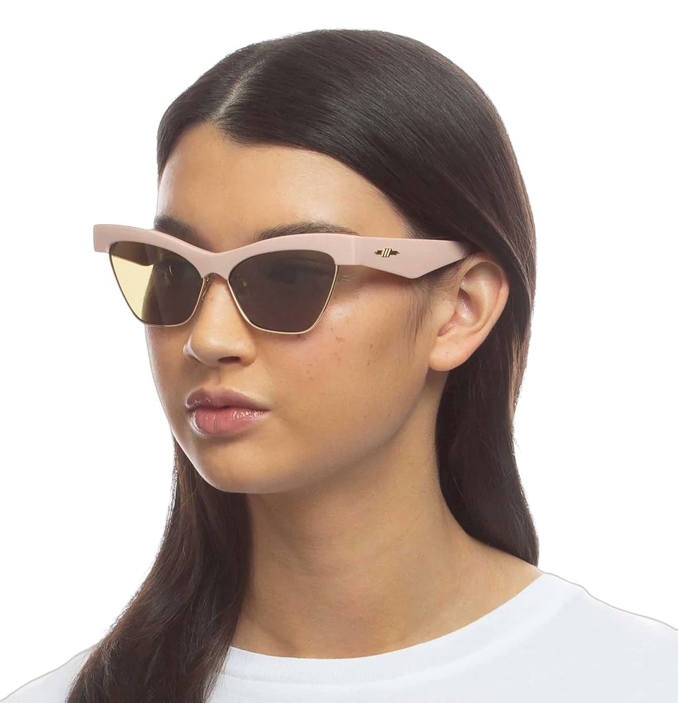 A woman wearing putty pink and gold Le Specs sunglasses with a cat-eye shape and metal details.